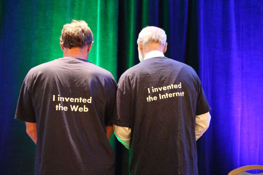 Tim Berners-Lee, inventor of the World Wide Web, stands with his back to the camera, wearing a t-shirt that says on the back "I invented the Web" and stands next to Vint Cerf, inventor of the Internet, who is wearing a t-shirt that says on the back "I invented the Internet."