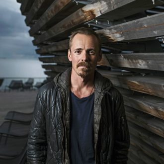 A portrait of Finnish actor Jasper Pääkkönen standing outside next to a wooden wall