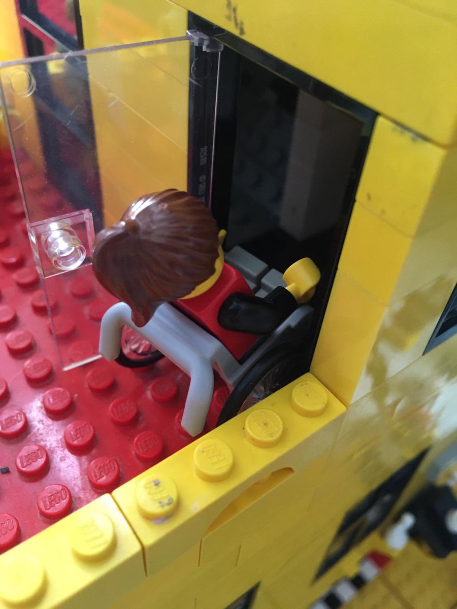 From above, a Lego figure with a brown pony tail tries to get her wheelchair through a door of a yellow and red building