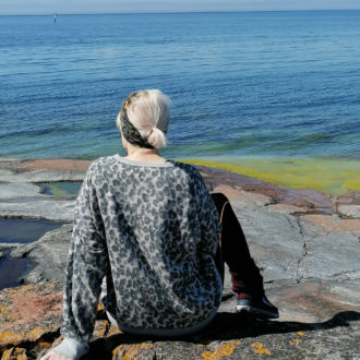 Dos tourné au photographe, une femme est assise sur une plage rocheuse, fixant le ciel et la mer s’étendant à perte de vue devant elle.