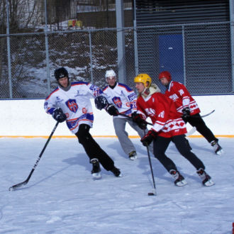 Cinq joueurs de hockey sur glace se pressent derrière le palet sur une patinoire en plein air.