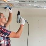 Young man in goggles fixing drywall suspended ceiling to metal frame using electrical screwdriver on ceiling insulated with shiny aluminum foil. Renovation, construction, do it yourself concept.