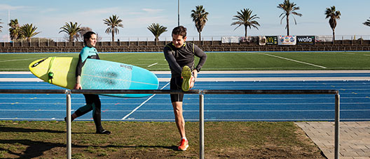 Una dona camina amb una planxa de surf i un home fa estiraments a la pista d'atletisme del Centre Esportiu Municipal La Mar Bella