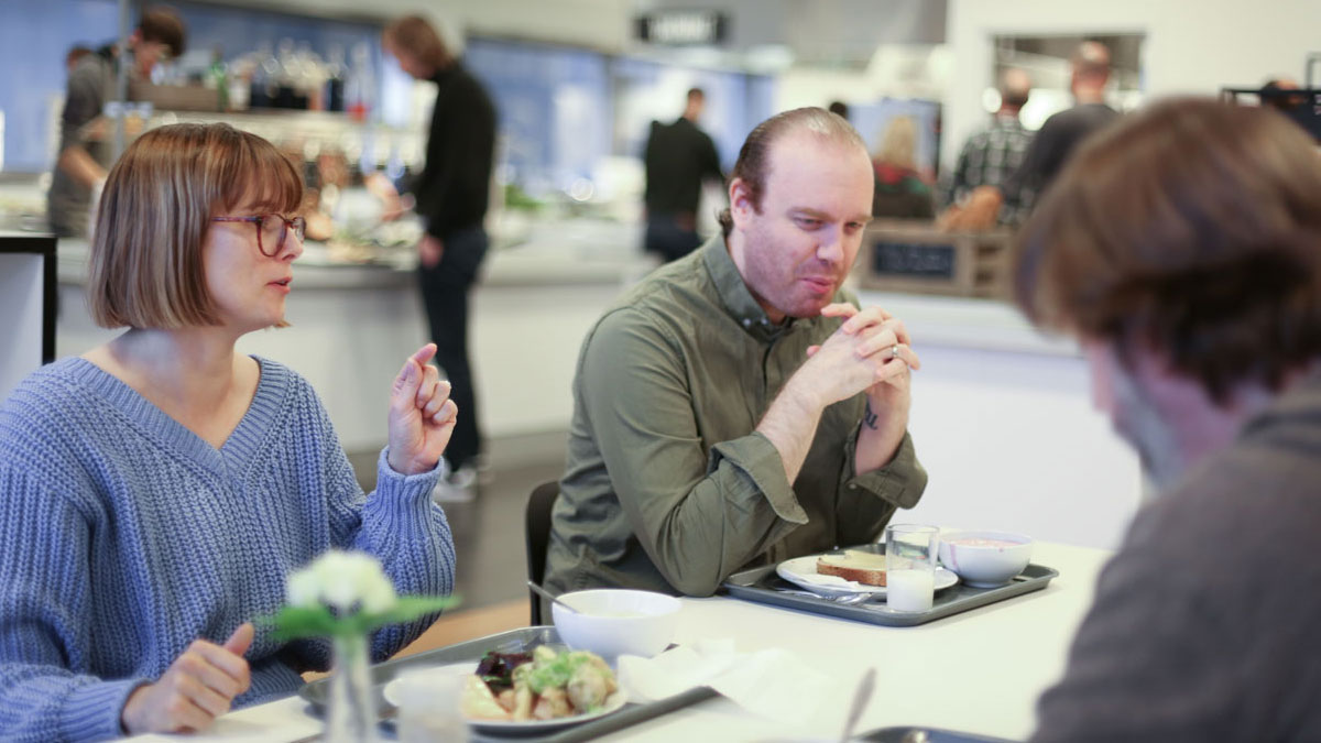 Drei Mitarbeiter unterhalten sich beim Mittagessen in einer Kantine.