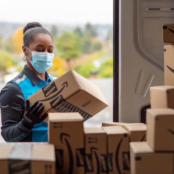 A woman wearing an Amazon delivery uniform picks up a package from the back of an Amazon truck full of packages. 