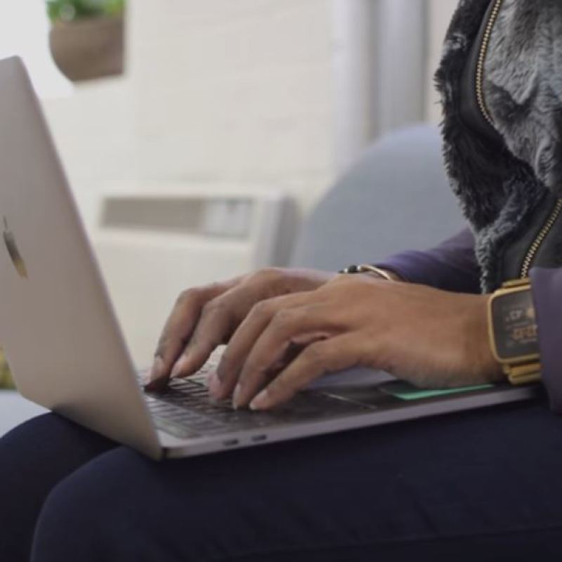 A woman types on the keyboard of a laptop computer, while sitting on a sofa. 