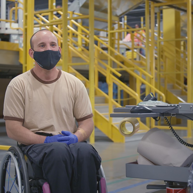 An Amazon Fulfillment Center employee wearing a black mask and protective blue gloves sits in a wheelchair next to a mobile computer desk.