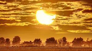 Picture with fields in the foreground and a crescent shaped Sun with Moon in front of it. Cloudy skies in the background.