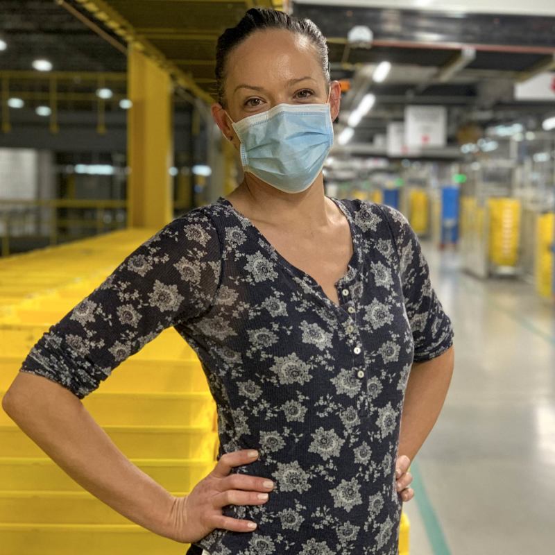 A woman wearing a mask stands in front of stacks of yellow plastic boxes.