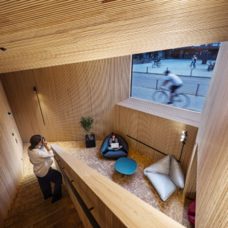 A woman sits in a pillow chair with a laptop, in a space where the walls, ceilings and stairs are all made out of wood.