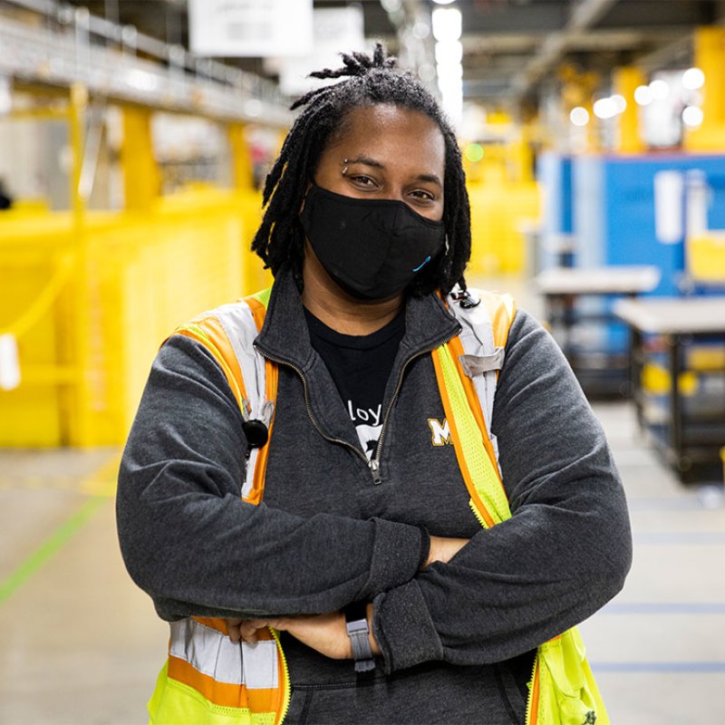 An Amazon employee stands in a fulfillment center with a mask on, looking at the camera.