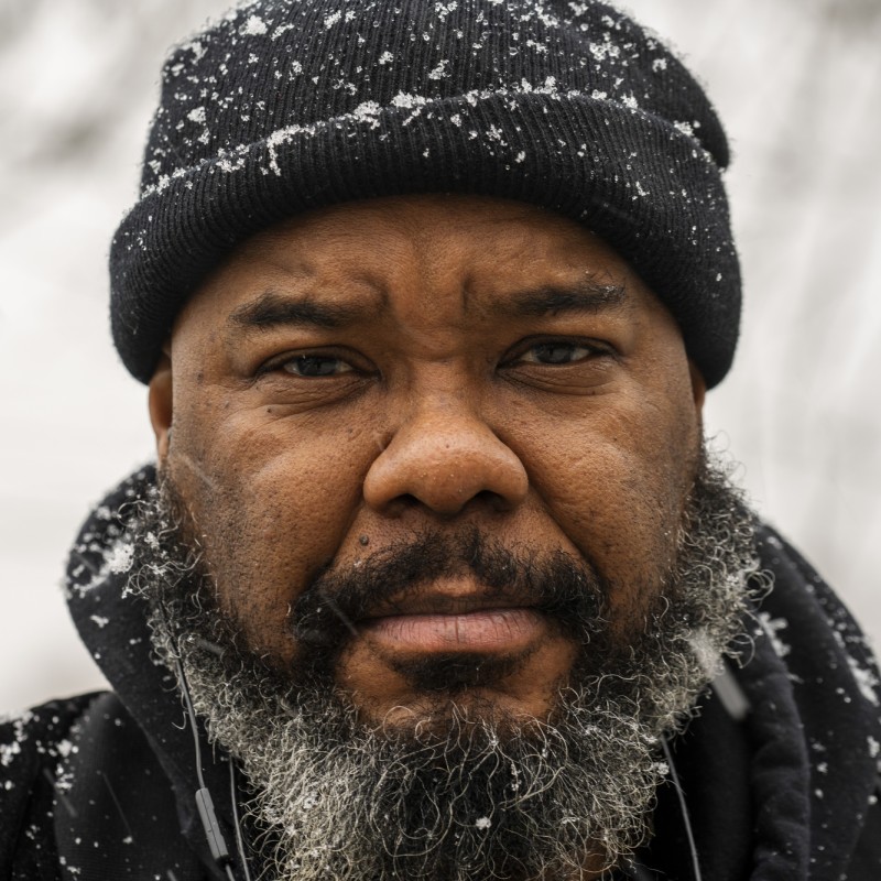 A bearded man in a winter hat stands outside in the snow.