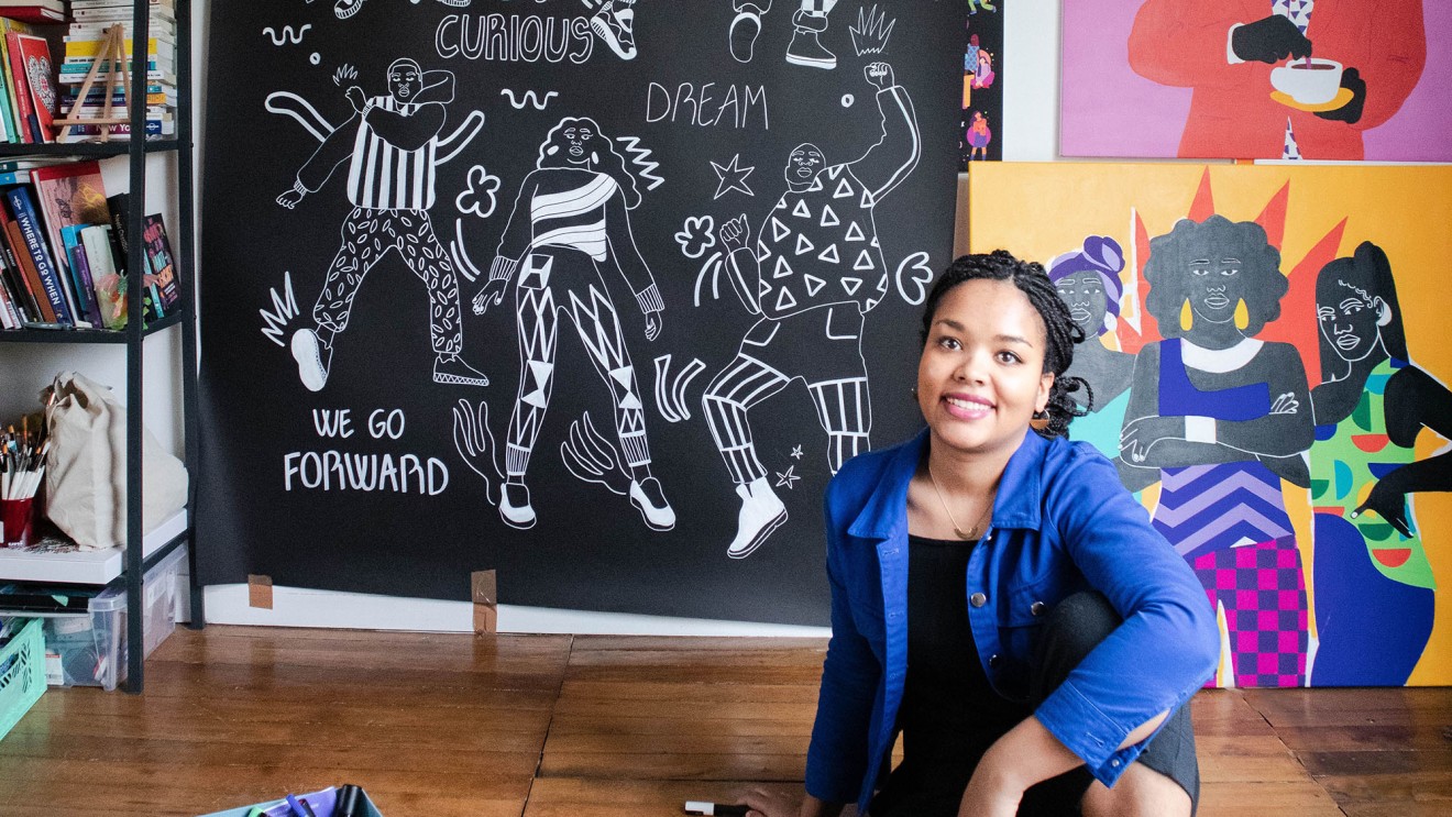 a Black woman, Aurelia Durand, sits on the wooden floor of her studio. Her artwork is displayed on the wall behind her, with saturated colors, and stylized people. 