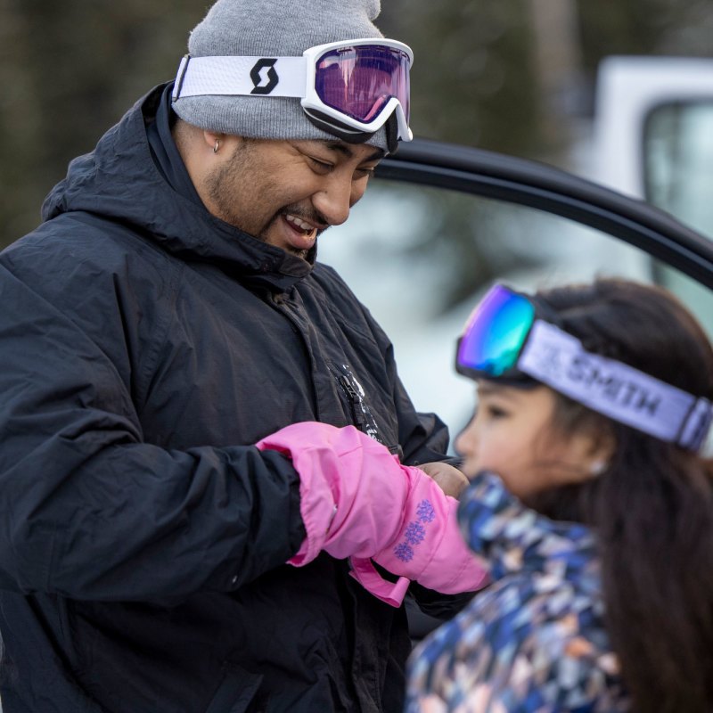 A father smiles as he looks down and helps his young daughter get her winter glove on.