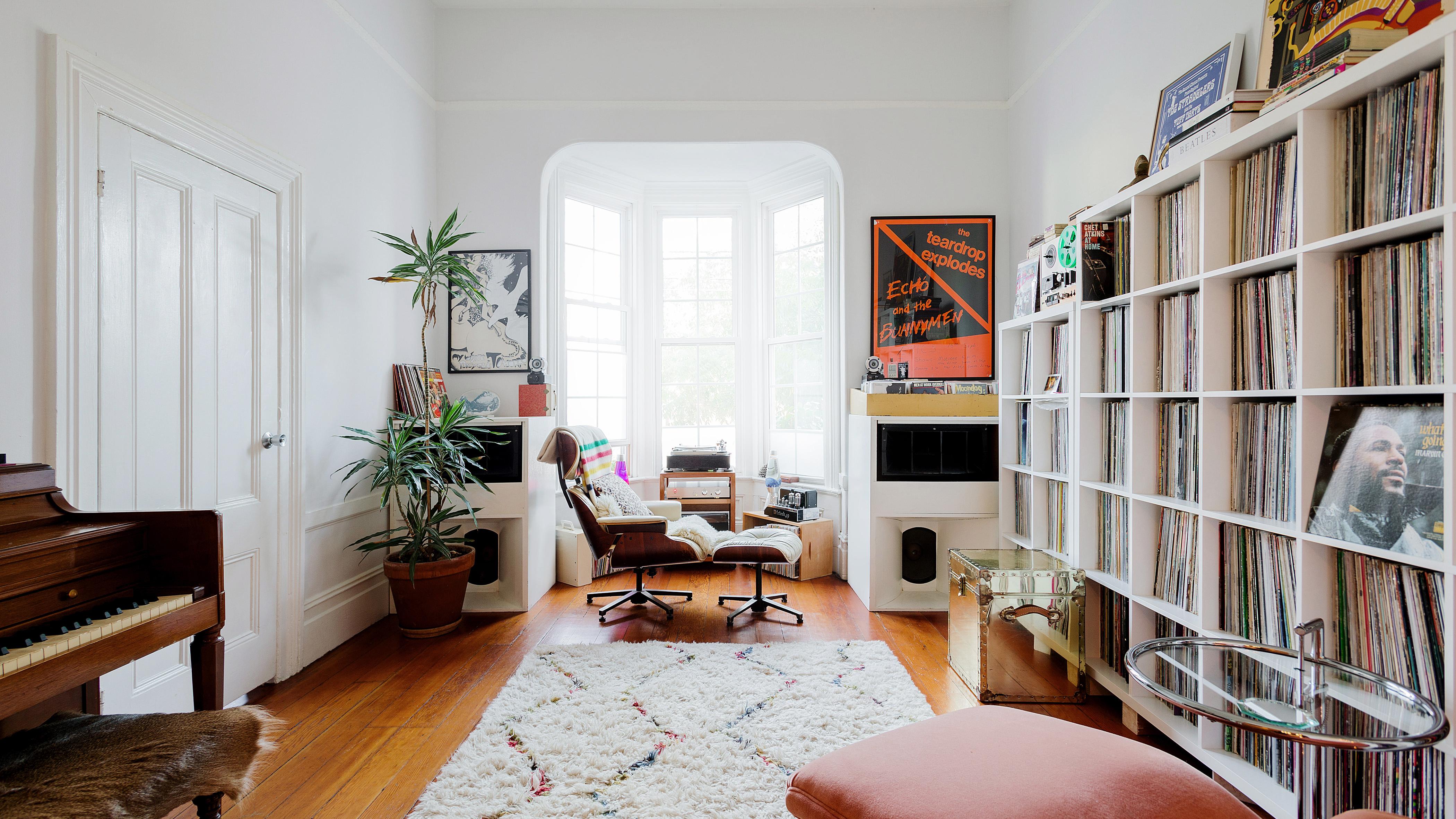 The bright living room, with a focal point on the large bay windows, record player, and a white leather and wood chair with matching ottoman. A large record collection spans the right wall, and on the opposite wall is an upright piano, door, and tropical plant.