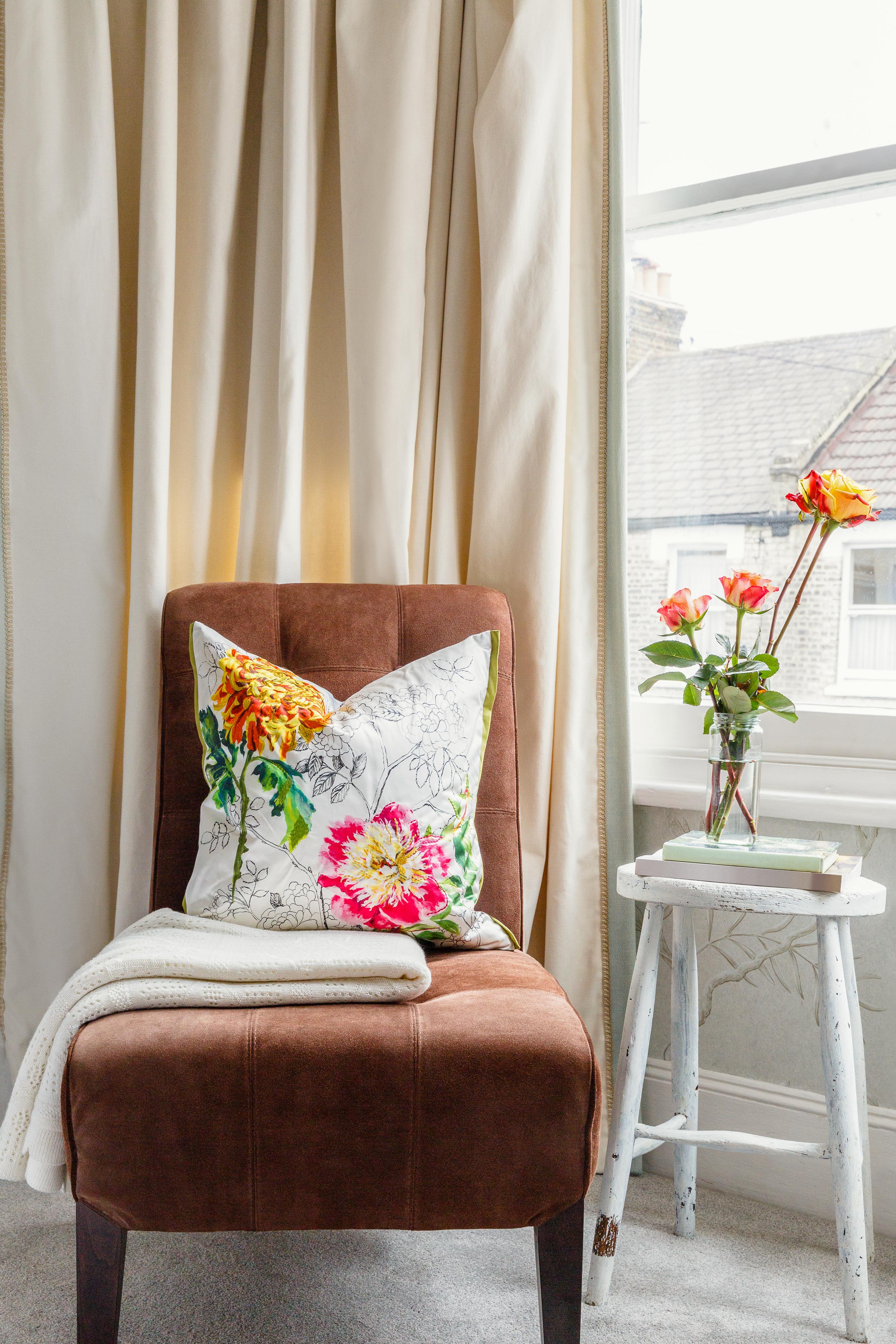 Window light fills a carpeted corner with drapes and a plush upholstered chair accented with a floral pillow and blanket. A case of fresh-cut flowers sits on top of a wooden side table.