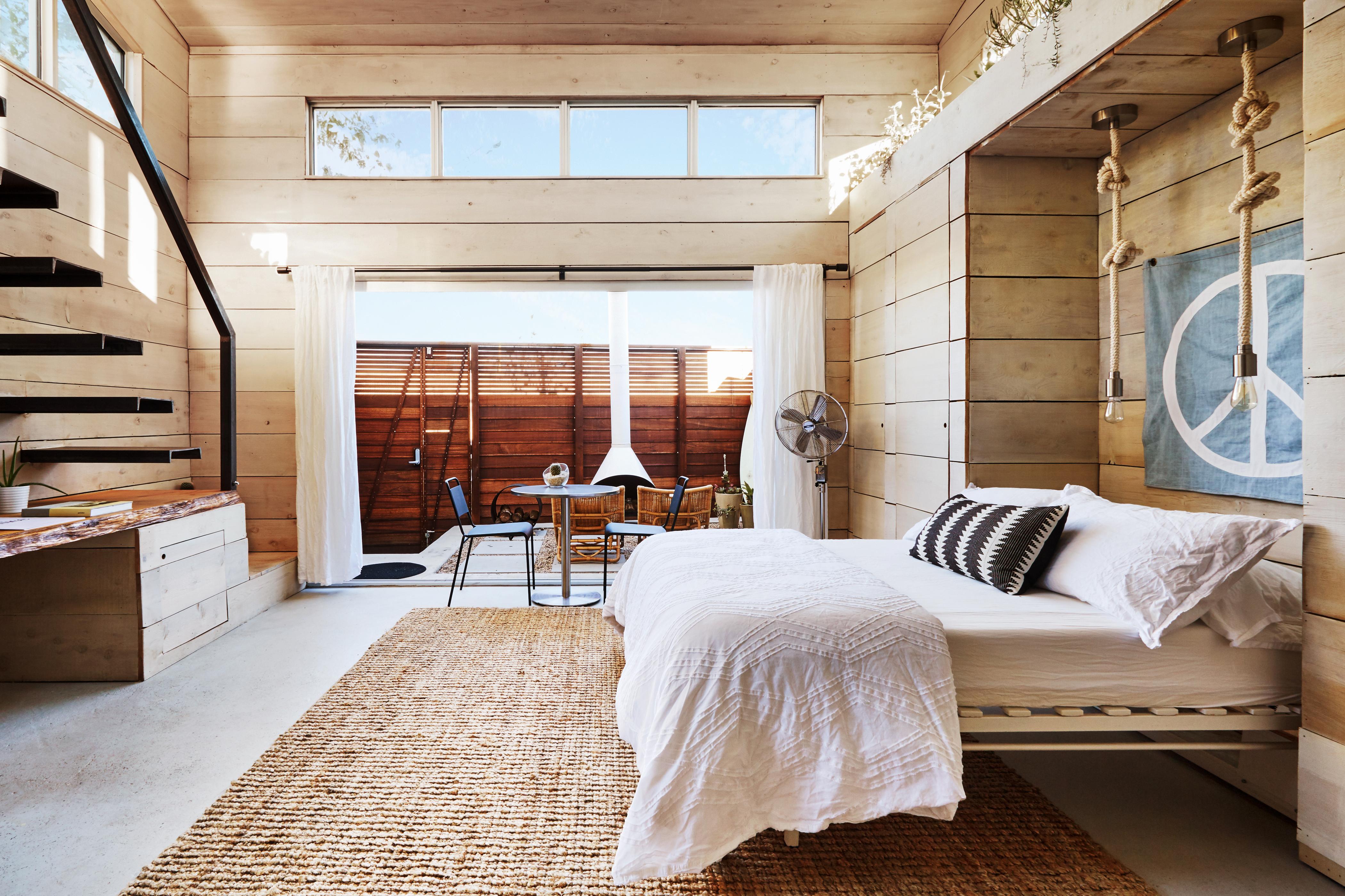 A spacious sleeping area with high ceilings, wood-slatted walls and windows. On the right is a bed, and to the left is a metal staircase that leads to what looks like a second level. The floor is covered by a large woven rug and at the back of room is a sliding glass wall that opens to a private patio with an outdoor fireplace.