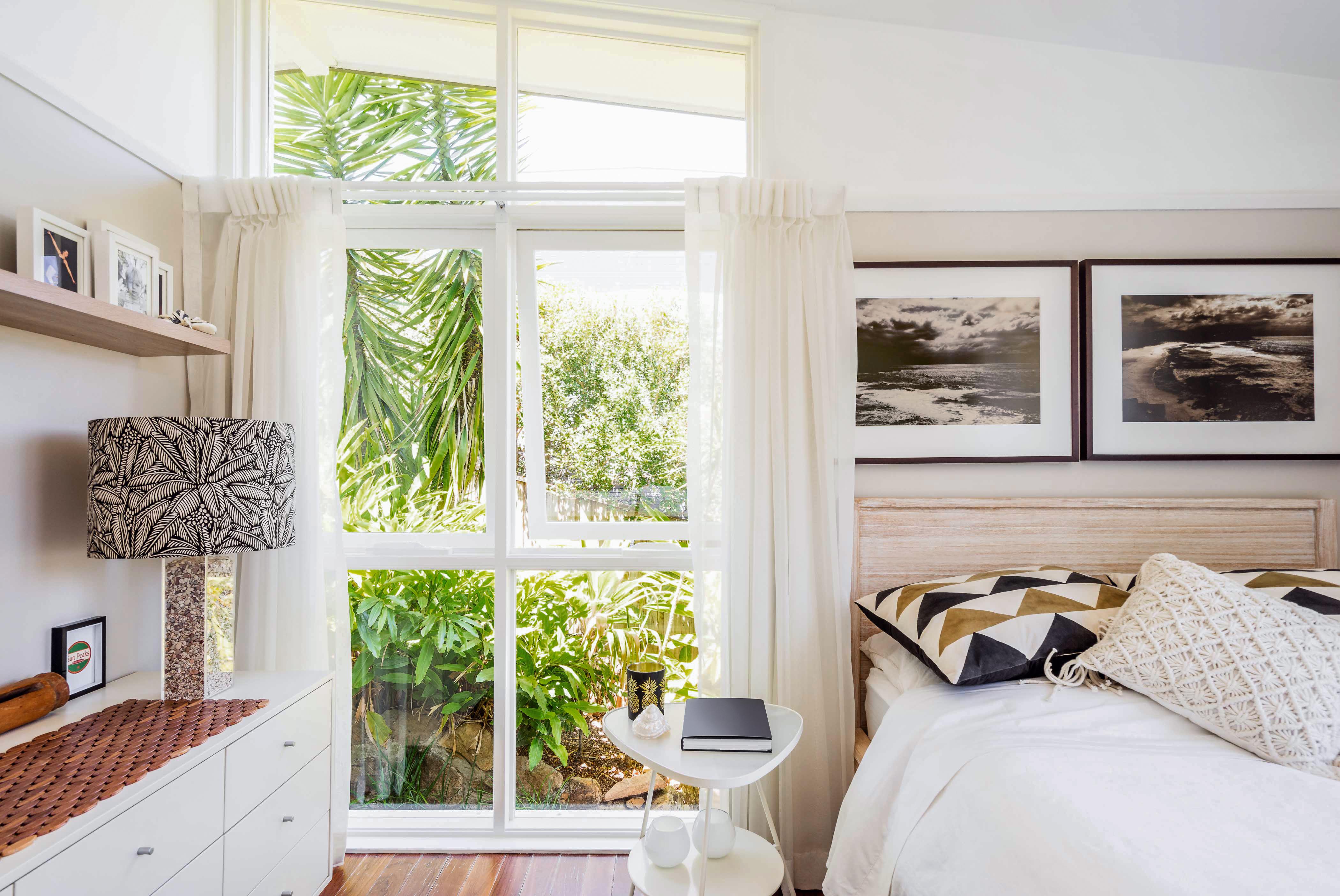 Bright corner of a bedroom with white walls, framed artwork, and a small bedside table. White drapes partially cover floor-to-ceiling windows, one pane of which is slightly opened.