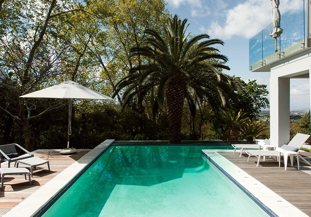 A modern, stone and glass home sits in a sunny landscape with a still, blue pool at its doorsteps.