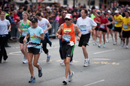 Older man and younger woman in running gear and bibs participating in the Boston Marathon near the one-mile remaining mark in Kenmore Square.