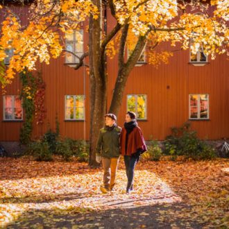 A man and a woman walking in front of red wooden houses and a fall foliage coloured tree and leaves