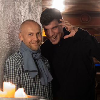 In a restaurant located in a cellar, in front of a table with lit candles on it, a man dressed in black makes a scary face at another man.