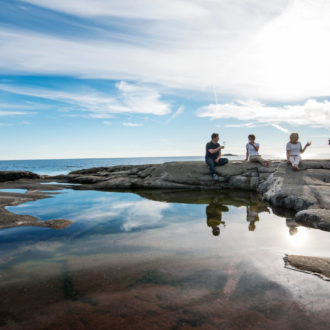 Four people sit on a large smooth rock by the ocean, their glasses raised in a toast.