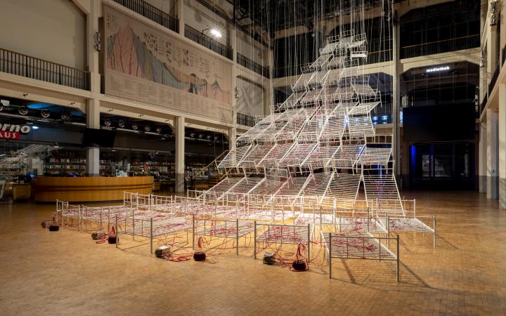 Installationsansicht von Chiharu Shiotas »Connected to Life«. Zu sehen sind mehrere Krankenhausbettgestelle, die von der Decke hängen. Durch transparente Schläuche an den Betten fließt rote Flüssigkeit.