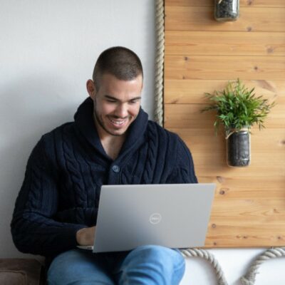 Man in blue jeans and hoodie using Dell Inspiron laptop on a bench with plants in the background.