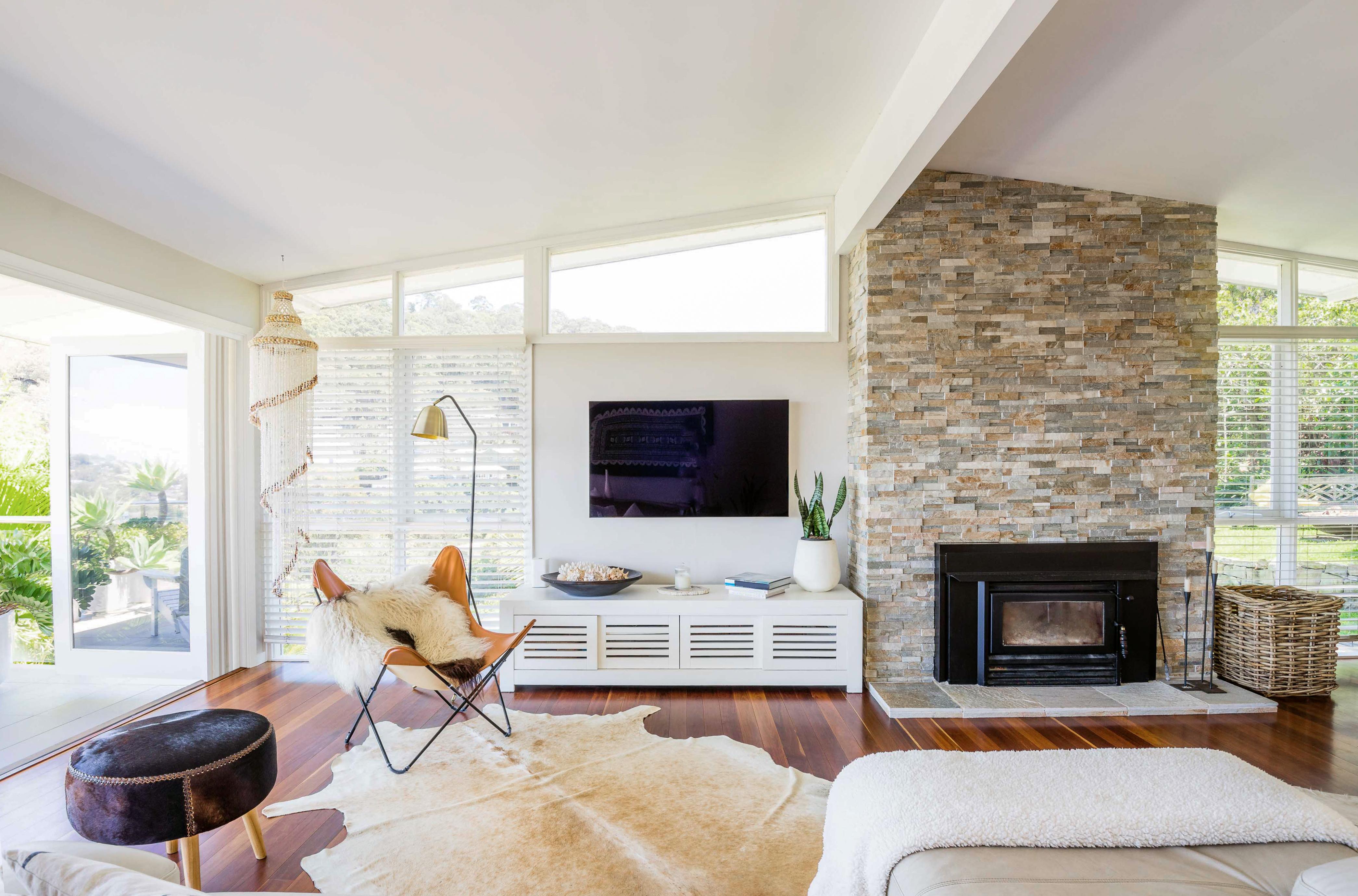 Light-filled, mid-century modern style living room with a vaulted ceiling, floor-to-ceiling windows, and a sliding glass door that opens to an outdoor area. In the background are windows, a stone fireplace, and a wall-mounted TV. The floor is covered by a cowhide rug and the room is furnished with a leather chair and ottoman.