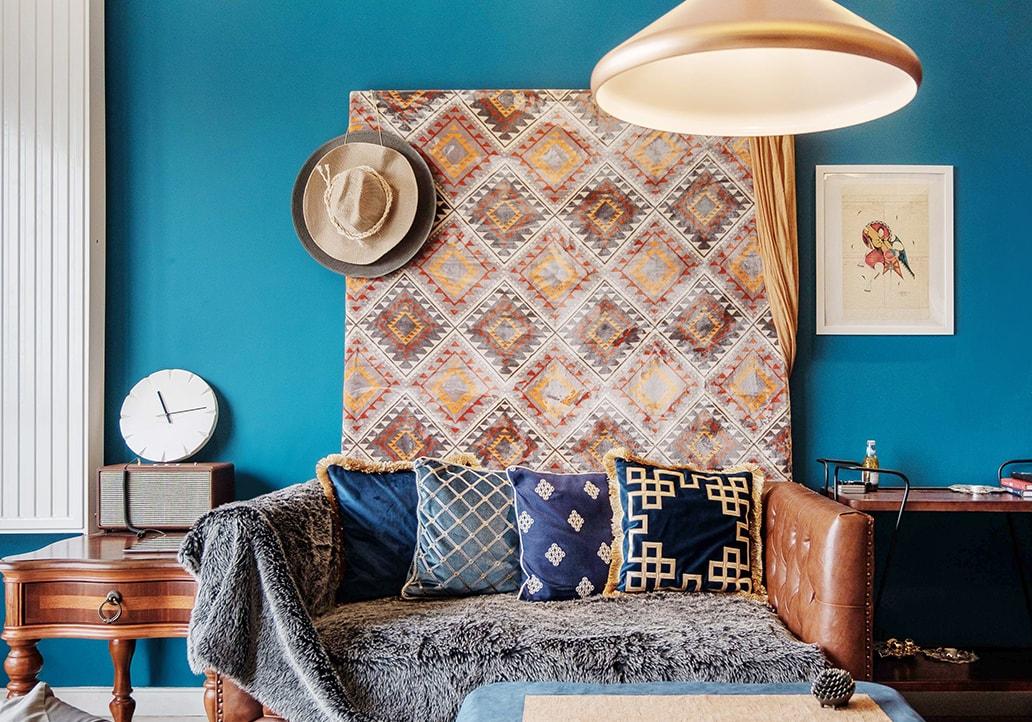 A brown leather couch with a soft blanket and square pillows sits in a blue room with rustic paintings and decorations on the walls.