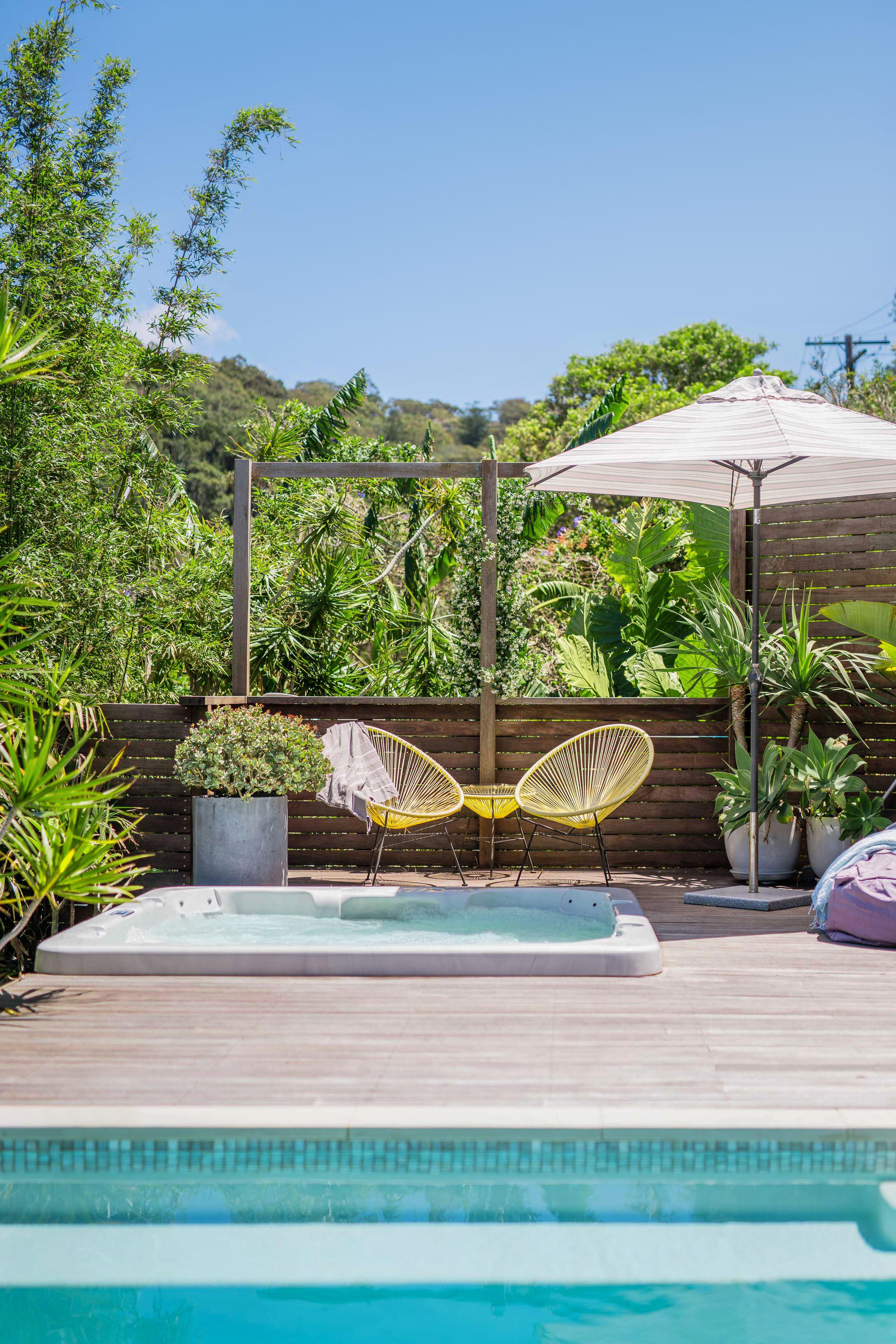 A wooden pool deck flanked by tropical plants and trees. Steps lead into a swimming pool partially lined in small turquoise tiles, and a hot tub built into the base of the deck is accompanied by two yellow chairs, a white umbrella, and potted plants.