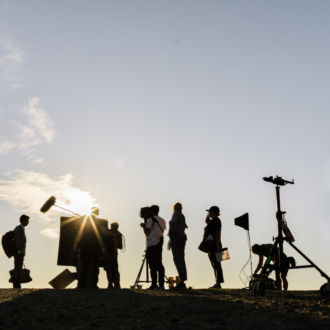 Silhouettes of people and camera equipment are next to a ship.