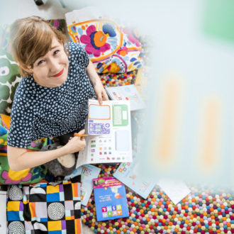 A smiling Linda Liukas surrounded with colourful pillows, computer stickers and children's books.