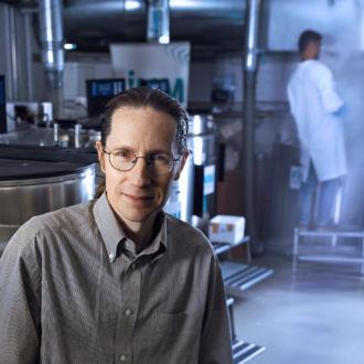 Un hombre con gafas de montura metálica y una camisa gris con el botón superior abierto, posa en un laboratorio científico.