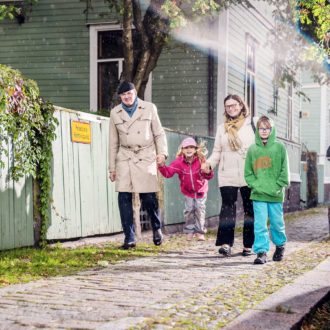 Walking past a wooden house, a man and a woman hold hands with a small girl who is jumping, while a boy walks beside them.