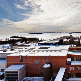 Under a covering of snow, a long brick building stretches out beside a bay, with islands visible in the distance.