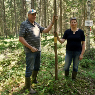 A man and a woman stand in the forest, each with one hand on a tall wooden pole that is stuck in the ground.