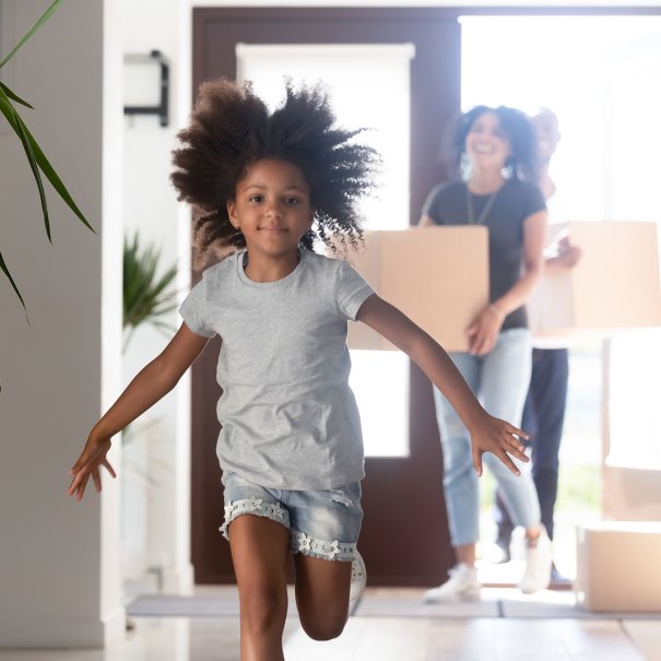 A little girl runs into a house.  Adults carrying moving boxes into the home are out of focus behind her.