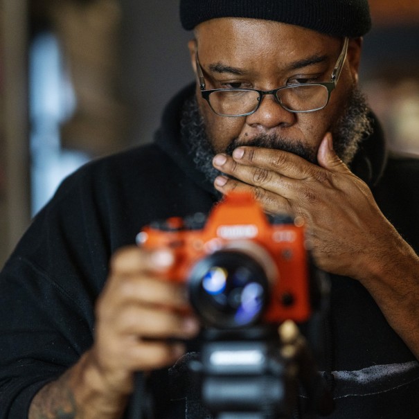 A man in glasses operates a camera mounted on a tripod.