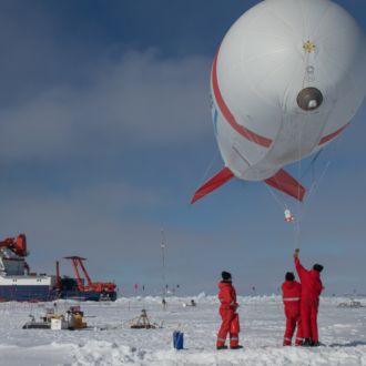 Three people in a snowy landscape getting ready to release a weather balloon, a ship in the background.