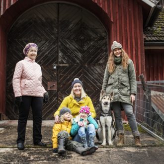 Eine ältere Frau, zwei Frauen in den Dreißigern und zwei kleine Kinder bekleidet mit Jacken und Mützen, die auf kühles Wetter hindeuten, posieren zusammen mit einem großen Hund vor einem Holzgebäude.