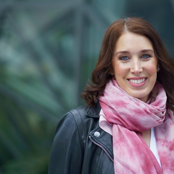 Sarah Gelman smiles for a photo in front of the Spheres at Amazon's headquarters in Seattle. She is wearing a pink scarf and a leather jacket.