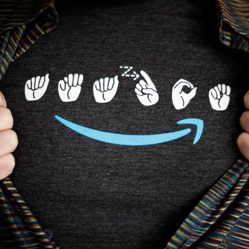 A man wears a t-shirt with the Amazon logo in sign language. Under the signs is a blue Amazon smile. The man is wearing a striped button down shirt over the t-shirt, and is holding the edges of his button down to make the entire logo visible.