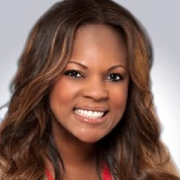 A woman who is Black smiles at the camera in front of a gray screen. 