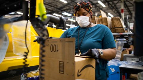 A woman working at an Amazon Fulfillment center wears a headwrap, a face mask, and gloves as she prepares a customer order. 