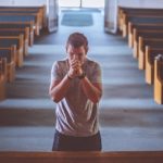 Ein Mann steht alleine vor dem Mittelgang einer Kirche Richtugn Altar in Gebetshaltung