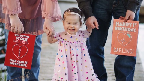 A toddler stands holding her mom and dad's hands. Her parents hold large greeting cards on either side of her. 