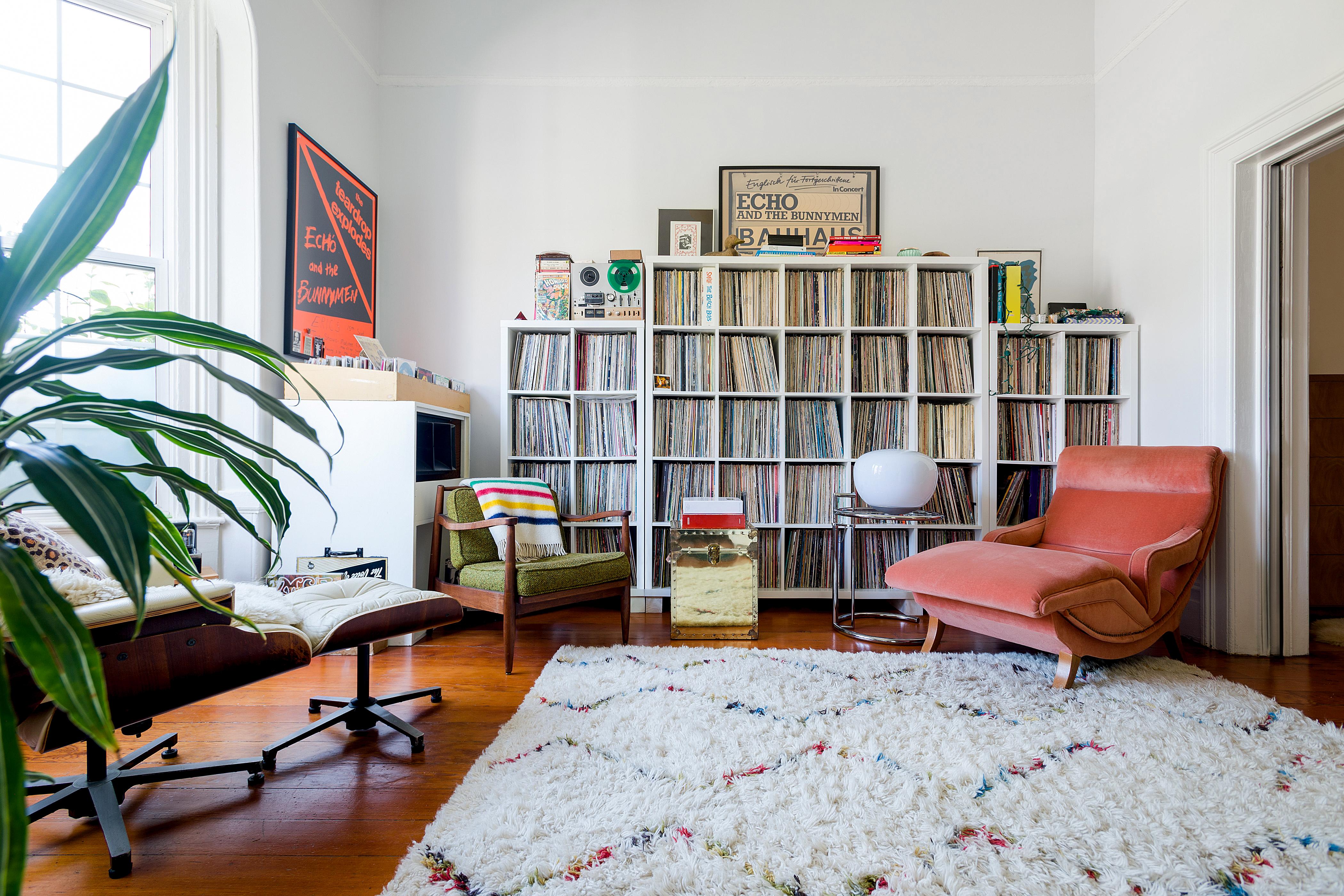 Ein Wohnzimmer mit Holzböden und hohen Fenstern. Der Blick wandert sofort zu einem texturierten Teppich und einer großen Plattensammlung an der hinteren Wand. Es gibt eingerahmte Musikposter und drei gepolsterte Sessel unterschiedlicher Größe und Textur.