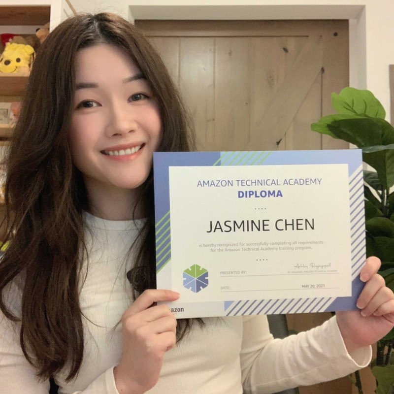 Jasmine poses with her ATA diploma. She has long dark hair wand wears a white long-sleeved shirt. There is a plant and drawings in frames behind her.